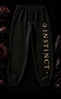Black jogger sweatpants with 'INSTINCT' printed vertically in gold on the left leg, displayed against a dark, textured background with gothic floral elements
