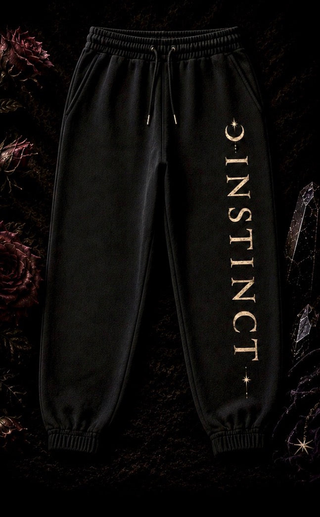 Black jogger sweatpants with 'INSTINCT' printed vertically in gold on the left leg, displayed against a dark, textured background with gothic floral elements